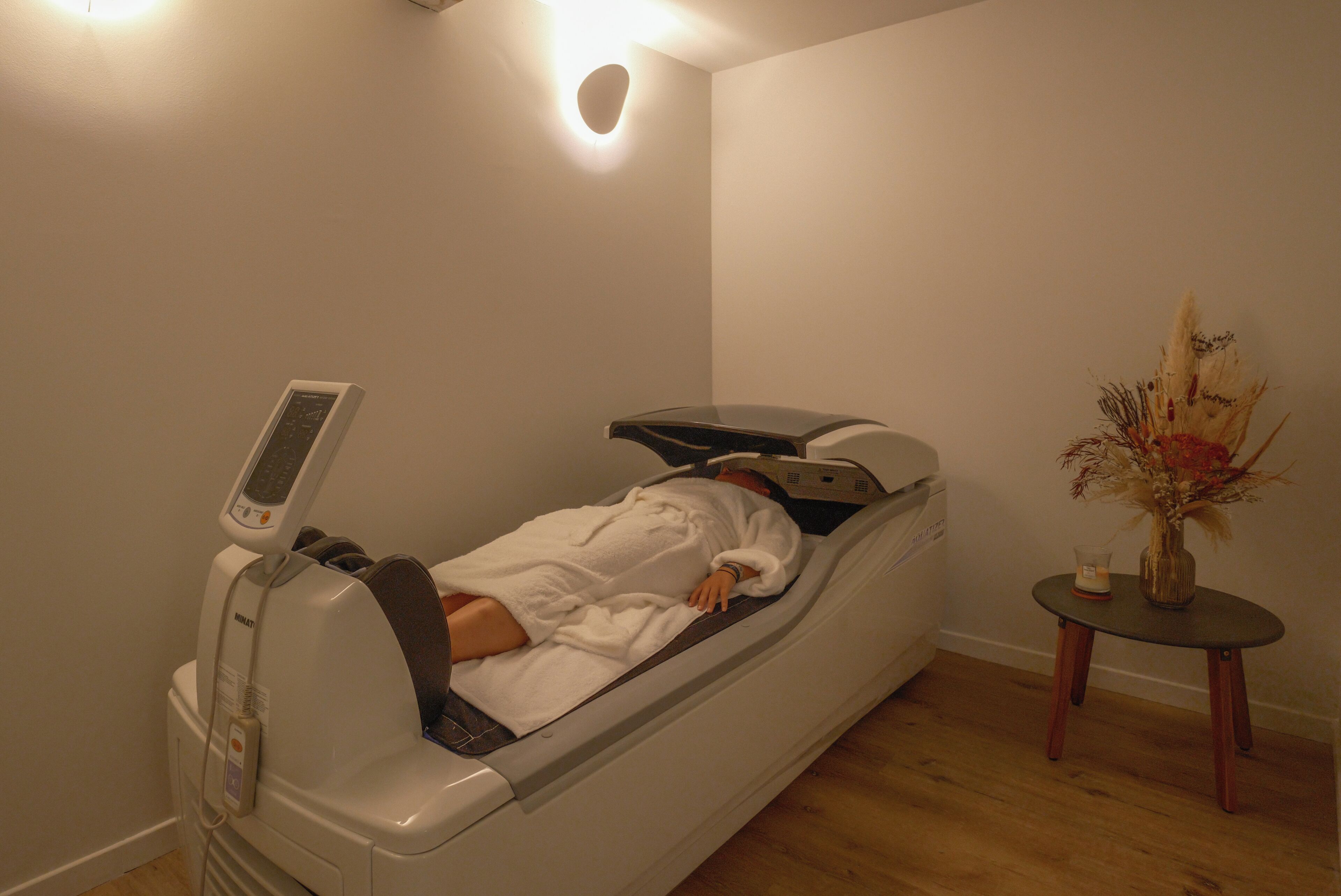 Un cuarto de terapia de masajes con una silla reclinable, un panel de control y una pequeña mesa con un florero.