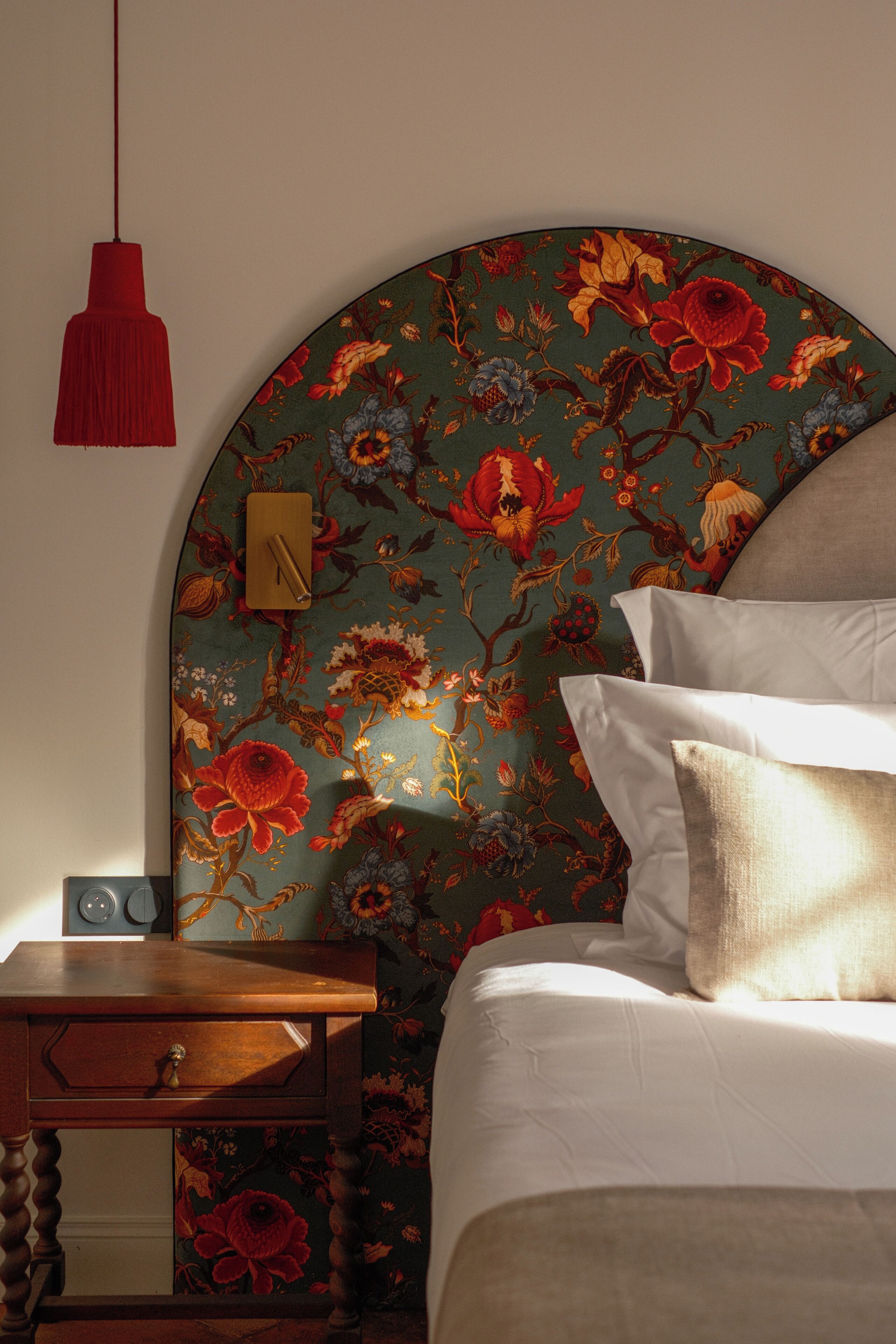 A bedroom with a floral wallpaper, a wooden nightstand, a red pendant light, and a bed with white bedding.