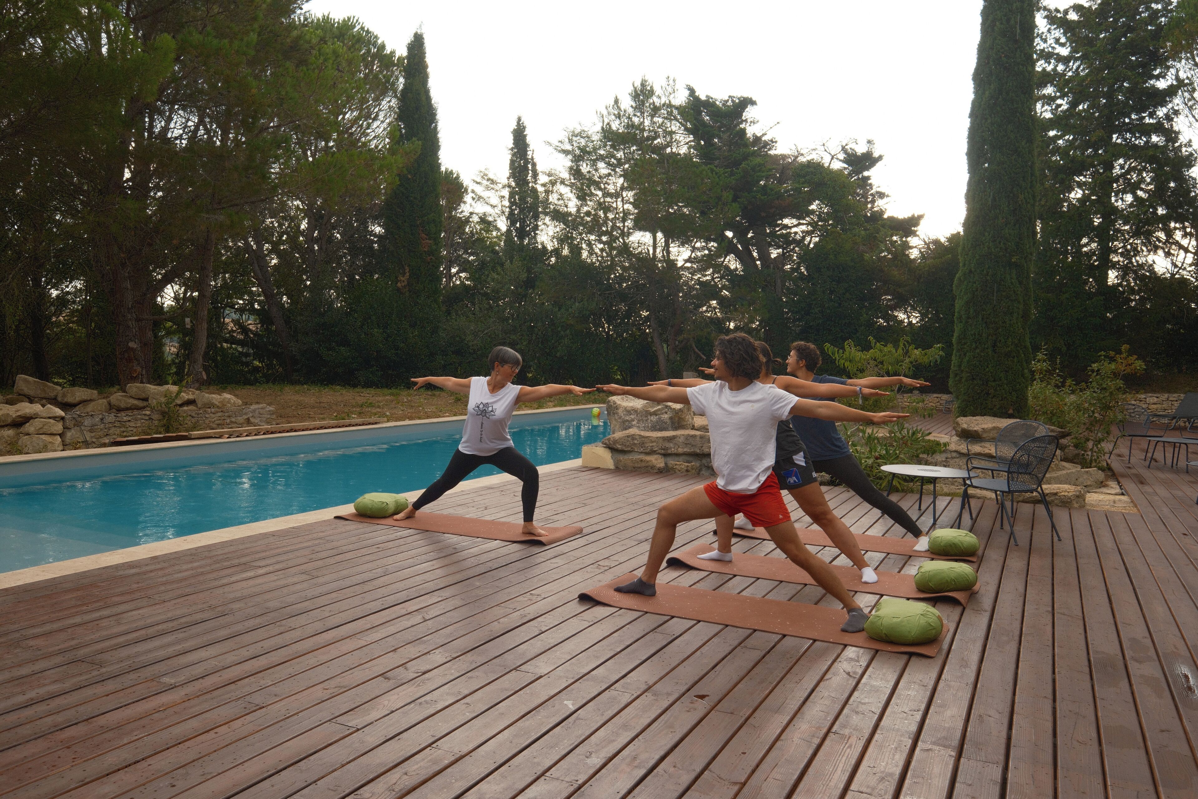 Un grupo de personas practicando yoga en una terraza de madera junto a una piscina, rodeado de árboles y mobiliario de exterior.