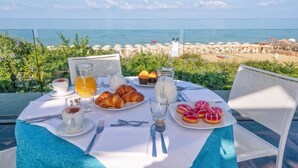 Une table dressée pour le petit-déjeuner, avec des croissants, des muffins et des viennoiseries, donnant sur une plage parsemée de parasols et de chaises longues.