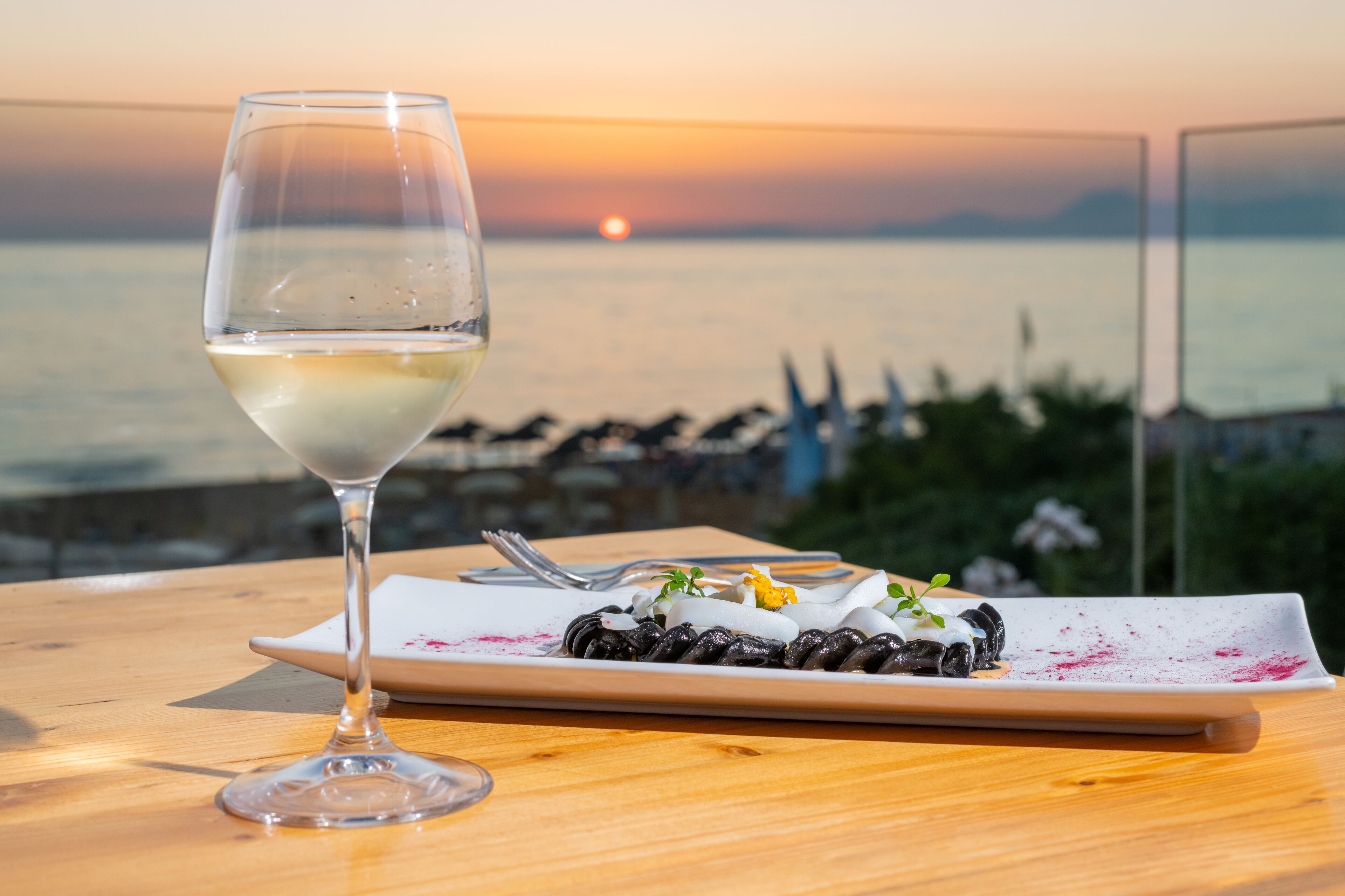 Una mesa con una copa de vino y un plato de comida, con vista a un cuerpo de agua al atardecer.