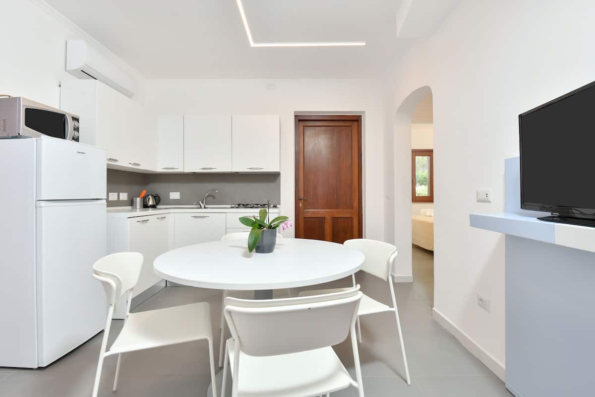 Une cuisine moderne avec des armoires blanches, un réfrigérateur blanc, un four à micro-ondes et une table à manger avec quatre chaises. On aperçoit une plante en pot sur la table et une porte en bois à l’arrière-plan.