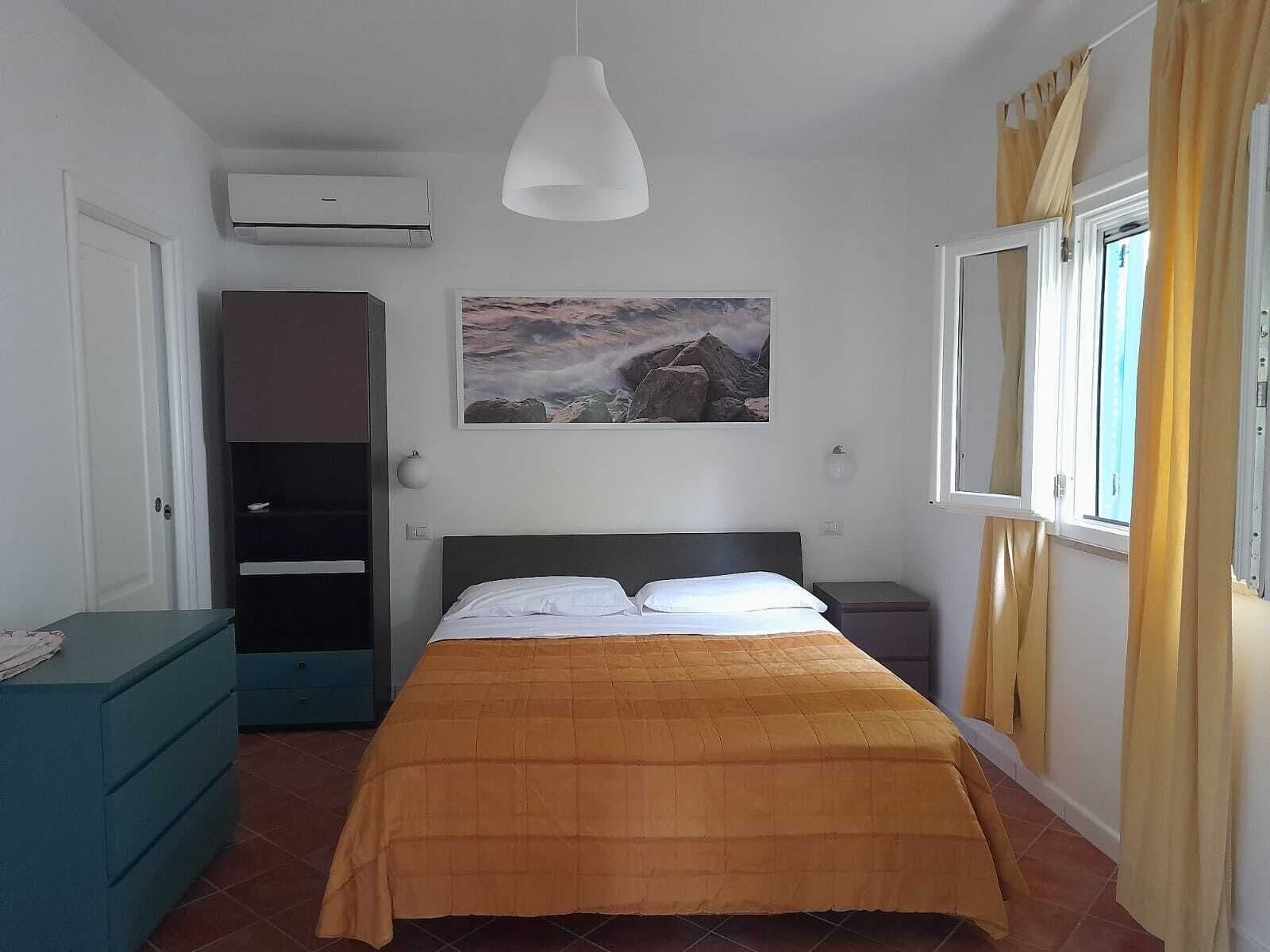 A bedroom with a bed, a wardrobe, an air conditioning unit, a painting, and a window with curtains.