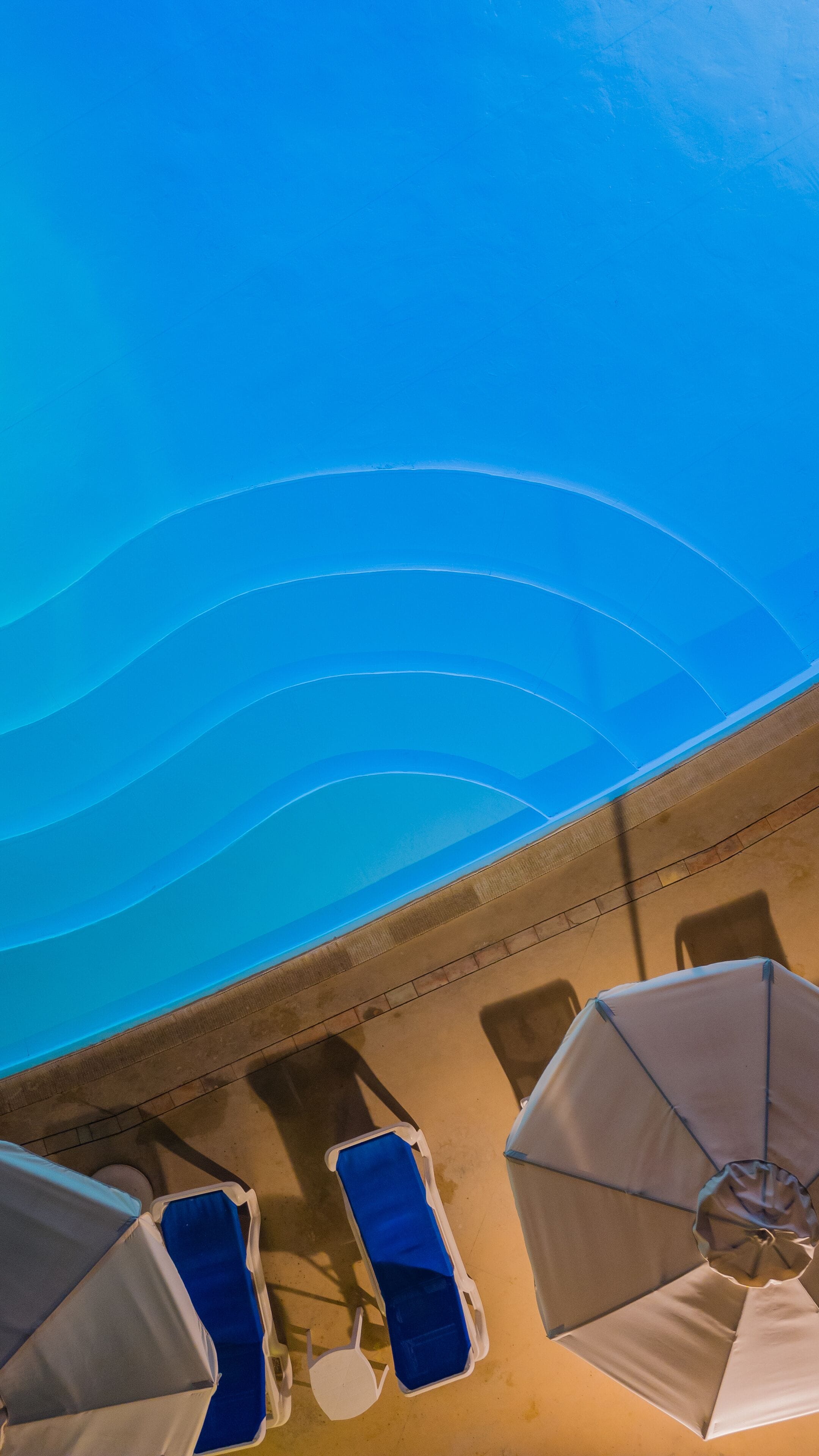 An aerial view of a pool area with blue lounge chairs and umbrellas, and a clear blue sky above.