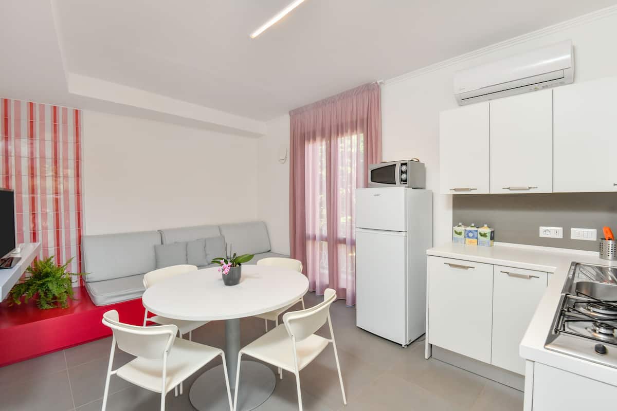 Une cuisine moderne avec des meubles blancs, un four à micro-ondes et une cuisinière à gaz. On y trouve un coin repas avec une table et des chaises blanches, ainsi qu’un banc rouge.