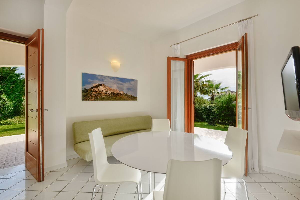Une pièce moderne avec une table ronde, des chaises blanches, un canapé vert et un tableau représentant un paysage.
