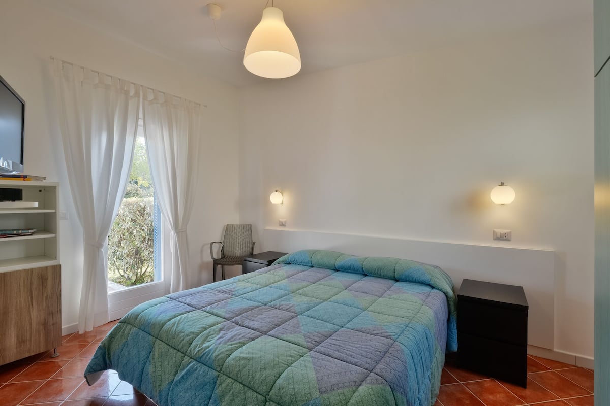 Une chambre à coucher avec un lit, une chaise, une table de chevet, une étagère et une fenêtre avec des rideaux.