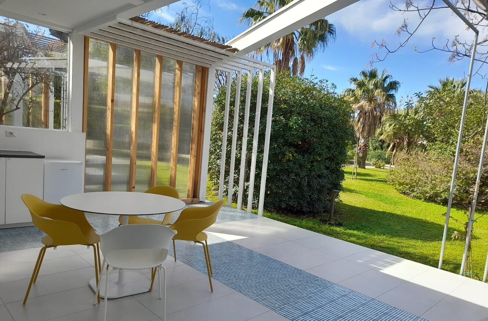 A modern outdoor dining area with a round table and yellow chairs, surrounded by a white pergola and a view of a well-maintained garden.
