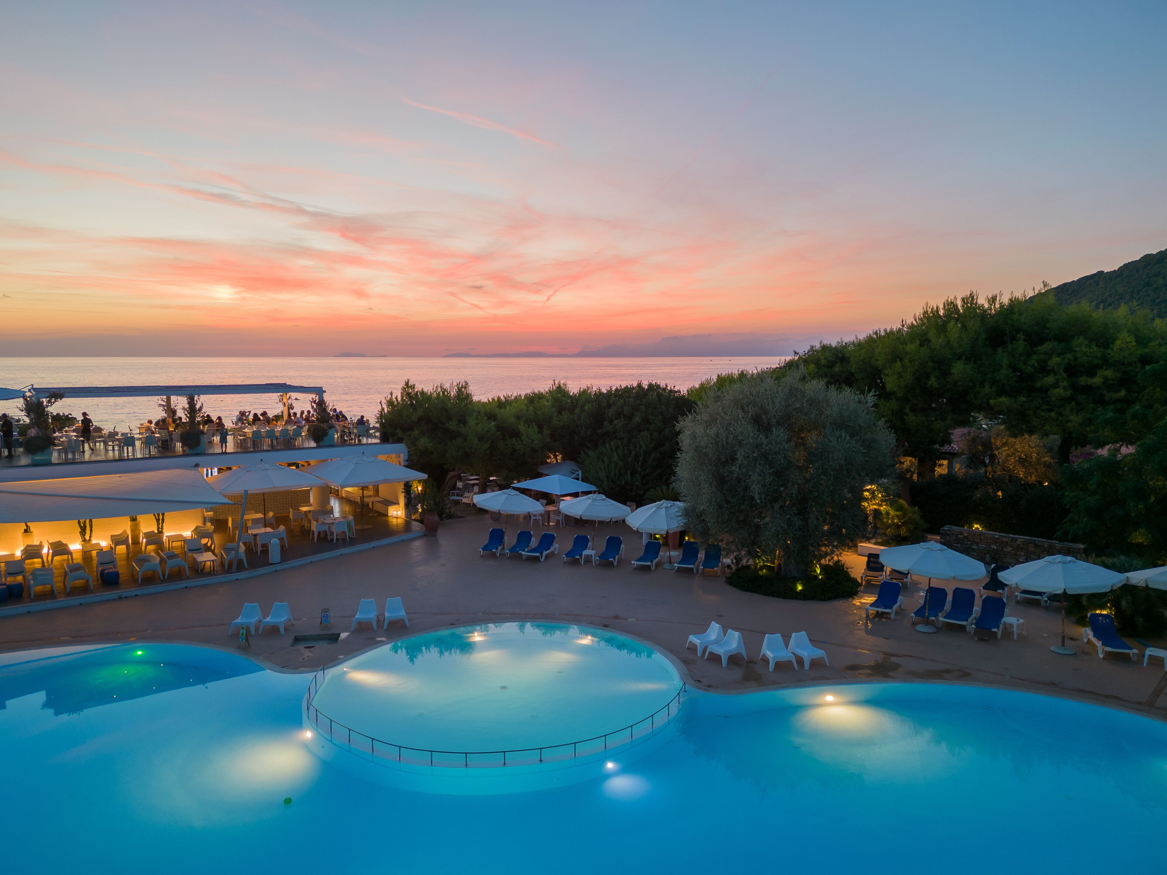 A resort pool area with lounge chairs and umbrellas, overlooking a body of water at sunset.