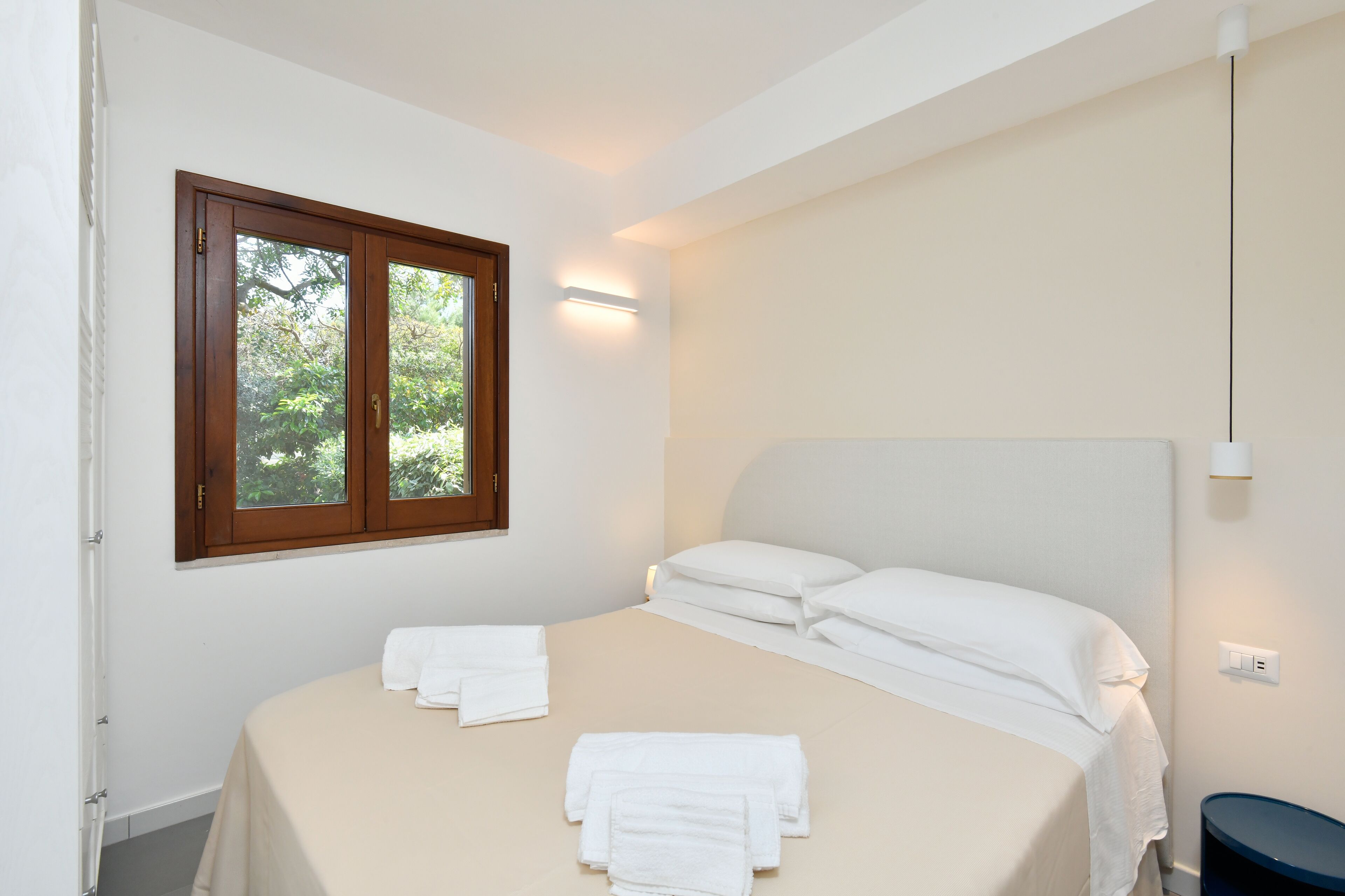 A bedroom with a bed, white linens, a window with a view of trees, and a wall-mounted light.