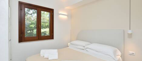 A bedroom with a bed, white linens, a window with a view of trees, and a wall-mounted light.