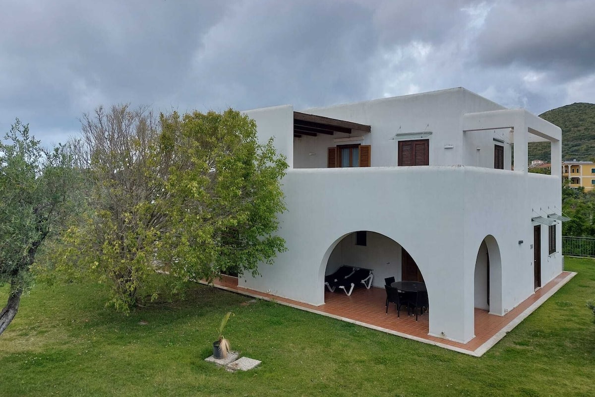 Une villa moderne, de couleur blanche, dotée d’un toit plat, de deux fenêtres apparentes et d’un petit patio aménagé avec du mobilier d’extérieur.