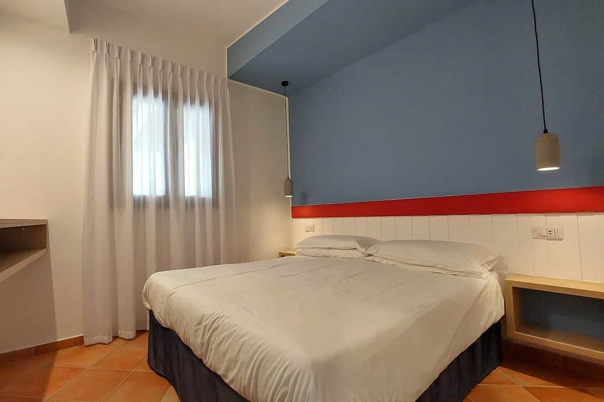 Une chambre à coucher comprenant un lit, une table de chevet, une étagère, une fenêtre avec des rideaux et un luminaire suspendu.
