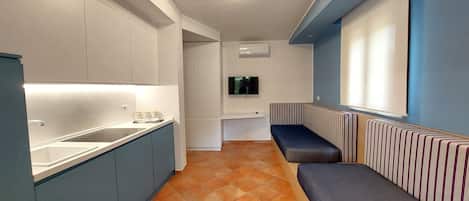 Una cocina compacta con estufa integrada, un lavamanos y una pequeña encimera. Hay un banco con cojines a rayas pegado a la pared, y un televisor de pantalla plana montado encima.