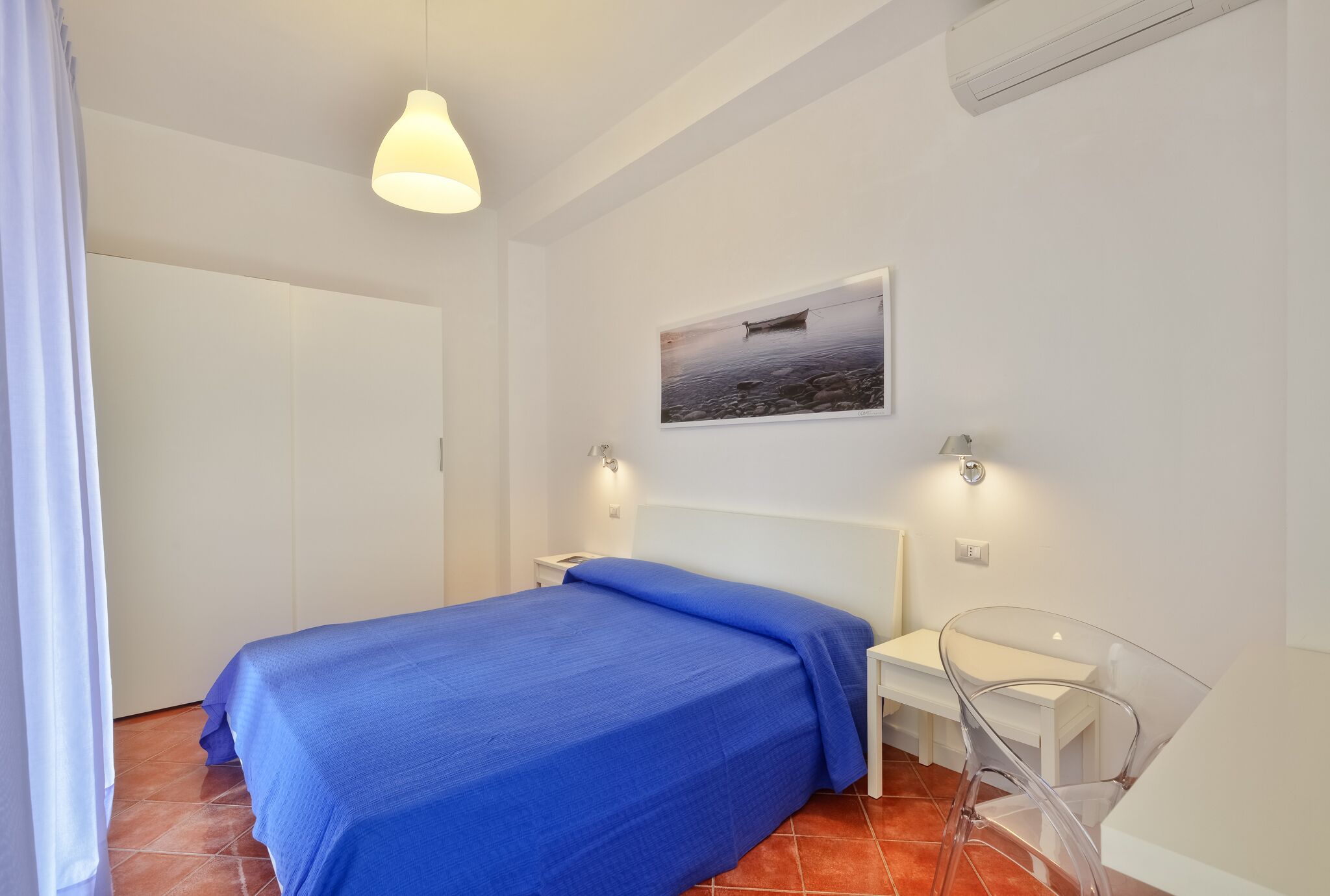 Een slaapkamer met een bed, nachtkastje, stoel, wanddecoratie en een plafondlamp.