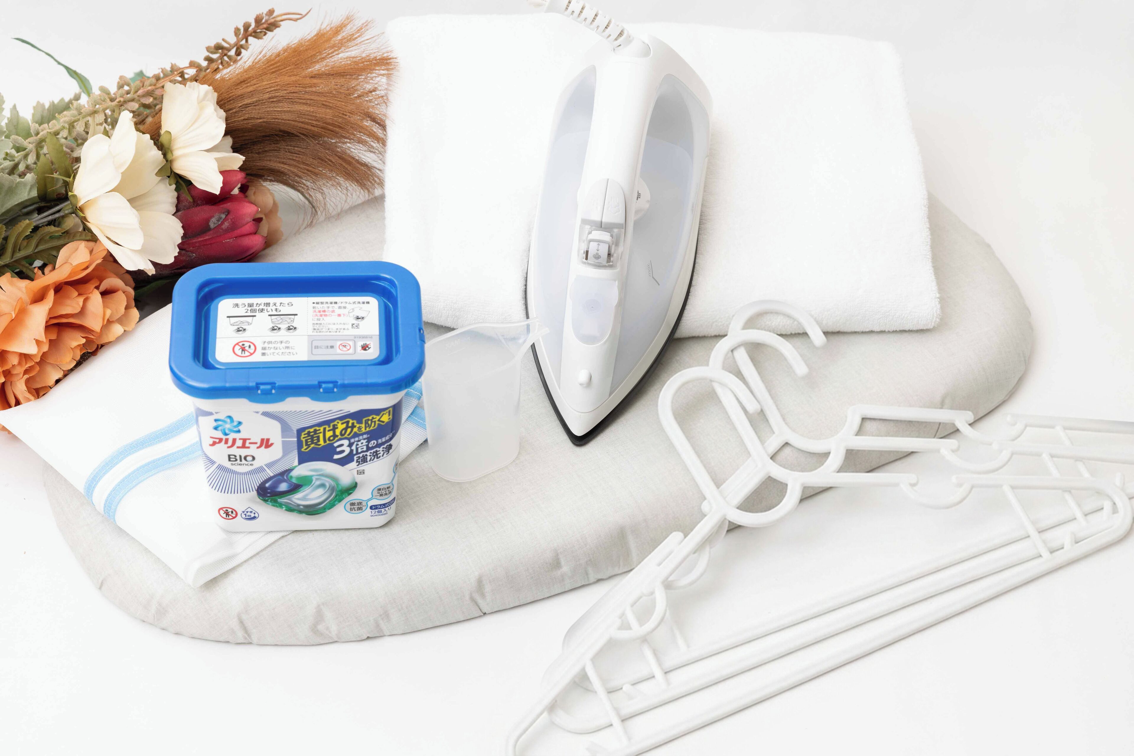 A white ironing board with an iron, a white towel, a blue container with a label, and a white hanger.