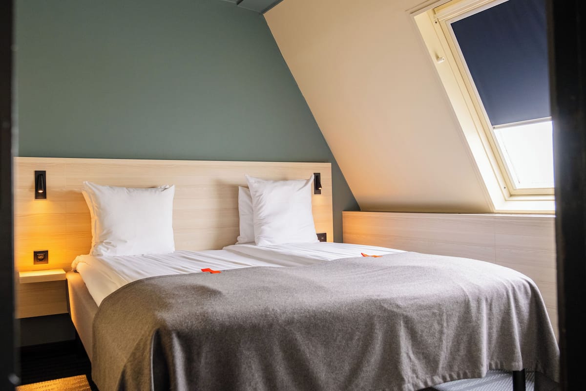 A bedroom with a sloped ceiling, a bed with white bedding, a bedside table with a lamp, and a window providing natural light.