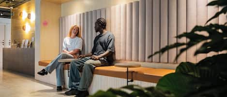 Dos personas sentadas en un banco en un vestíbulo moderno con un mostrador de recepción de madera y un panel de pared vertical.