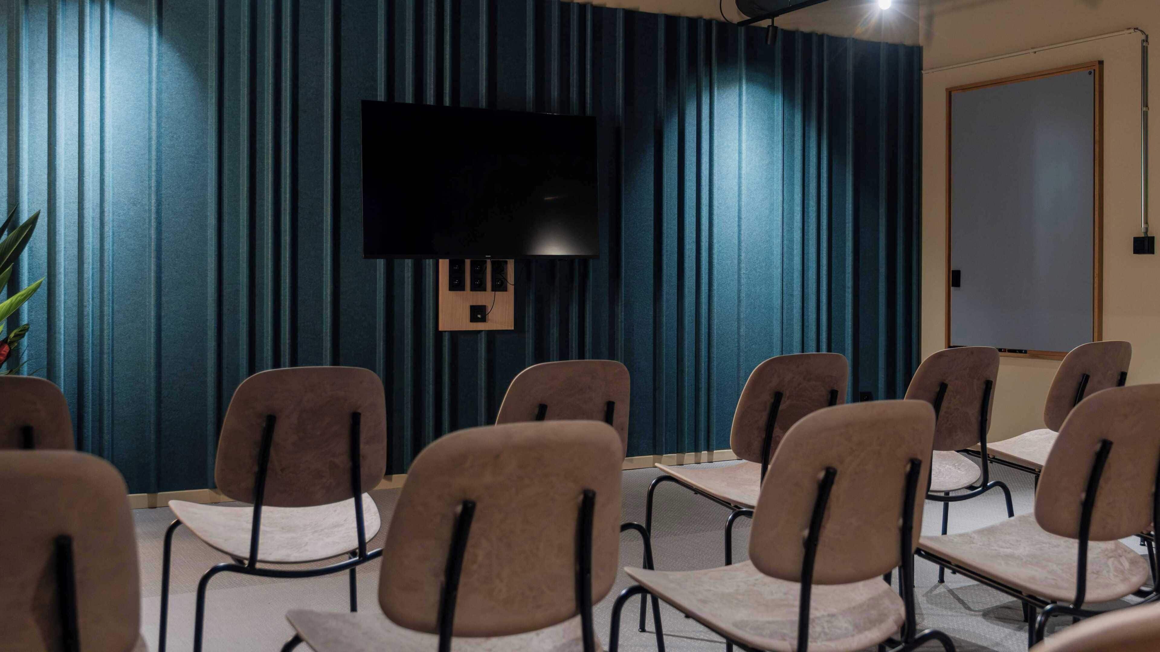 Ein Konferenzraum mit einem an der Wand befestigten Flachbildfernseher, einer Whiteboard-Tafel und mehreren Stühlen, die um einen Tisch angeordnet sind.