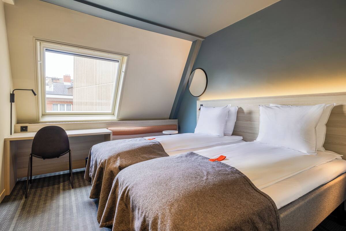A hotel room with two beds, a desk, a chair, and a window.