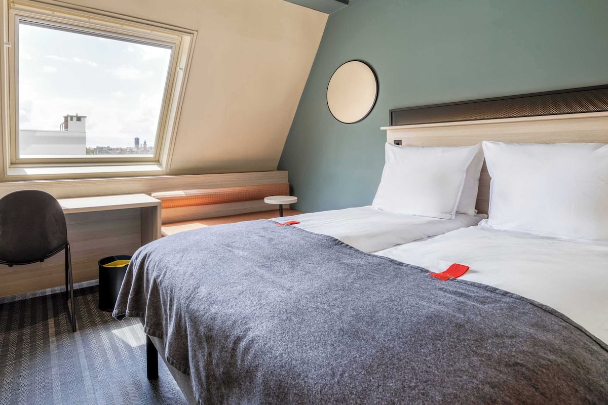 A hotel room with a bed, a desk, a chair, a round mirror, and a window with a city view.