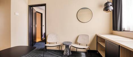 A hotel room with a desk, two chairs, a small round table, a mirror, a lamp, and a window with curtains.
