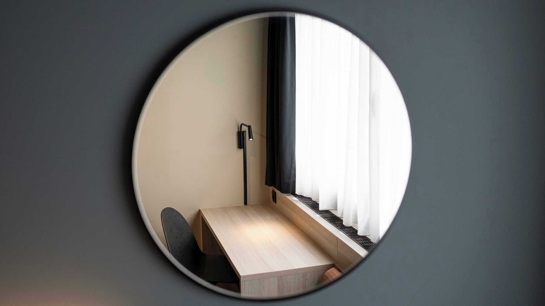 Un espejo circular refleja una esquina de una habitación con un escritorio, una silla, una lámpara de pie y una ventana con cortinas.