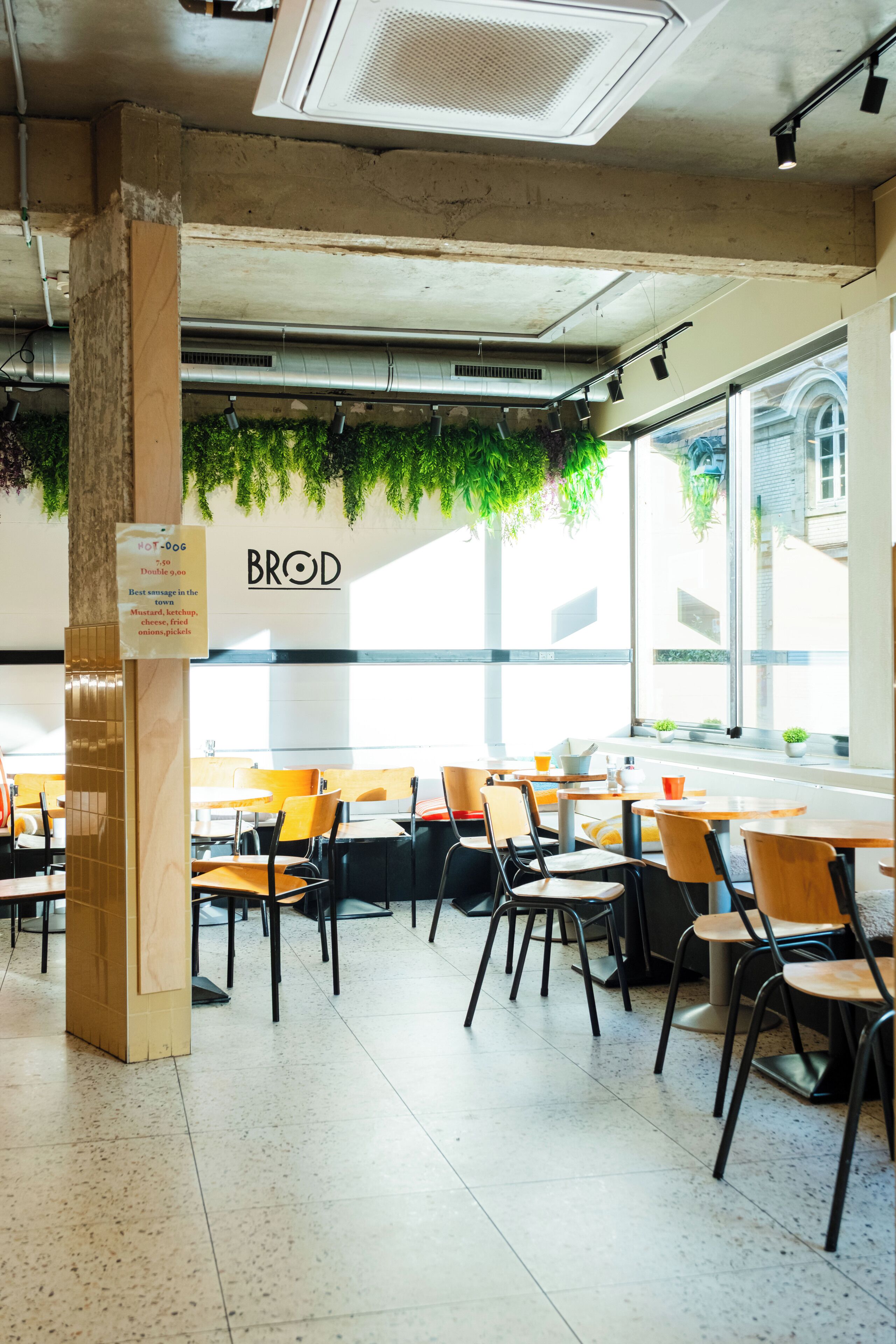 A restaurant interior with a high ceiling, exposed ductwork, and a menu board displaying the name 'BROD'. Tables and chairs are arranged for dining.