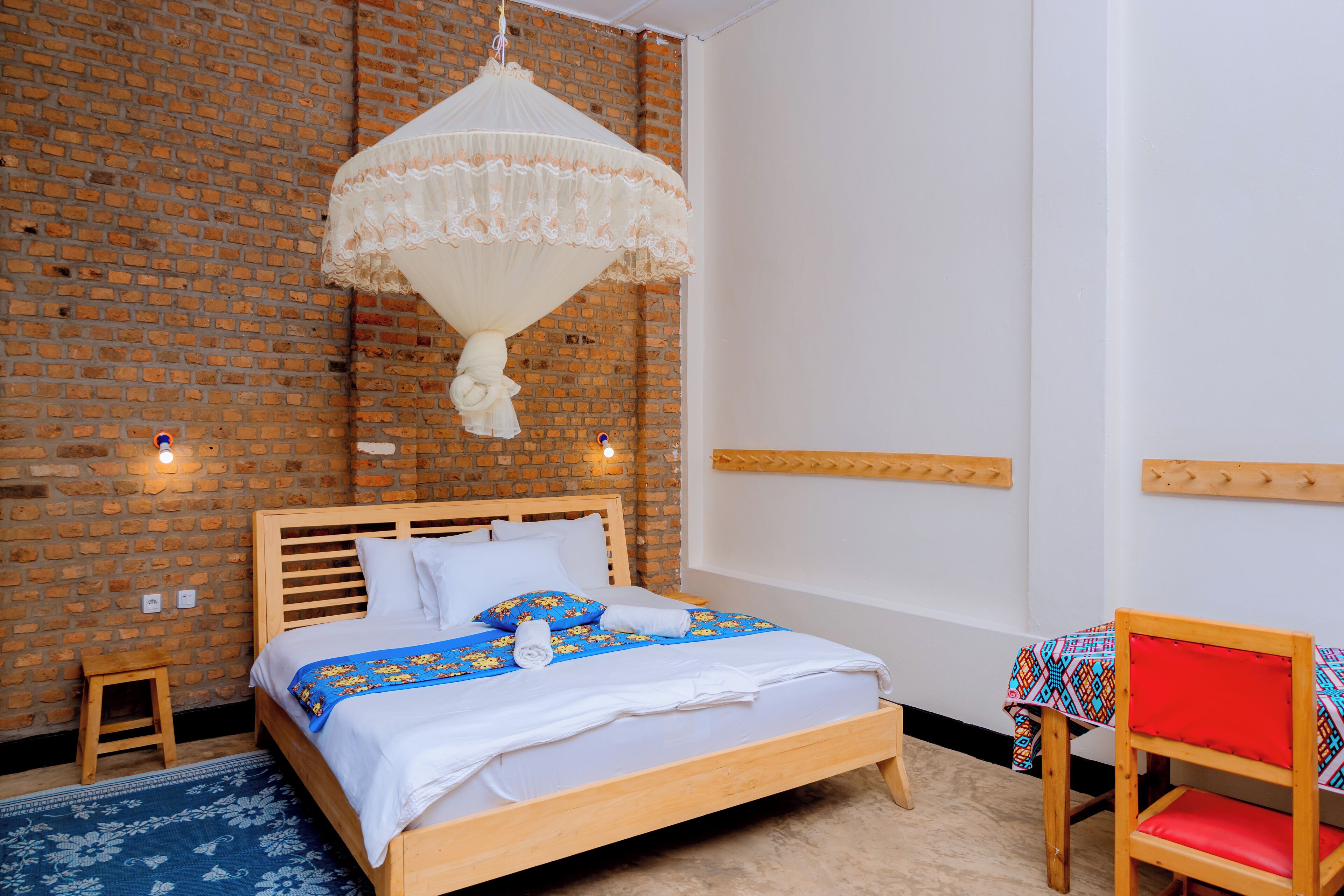 Un dormitorio con pared de ladrillo, una cama de madera, un dosel colgante, una mesita con mantel colorido y una silla roja.