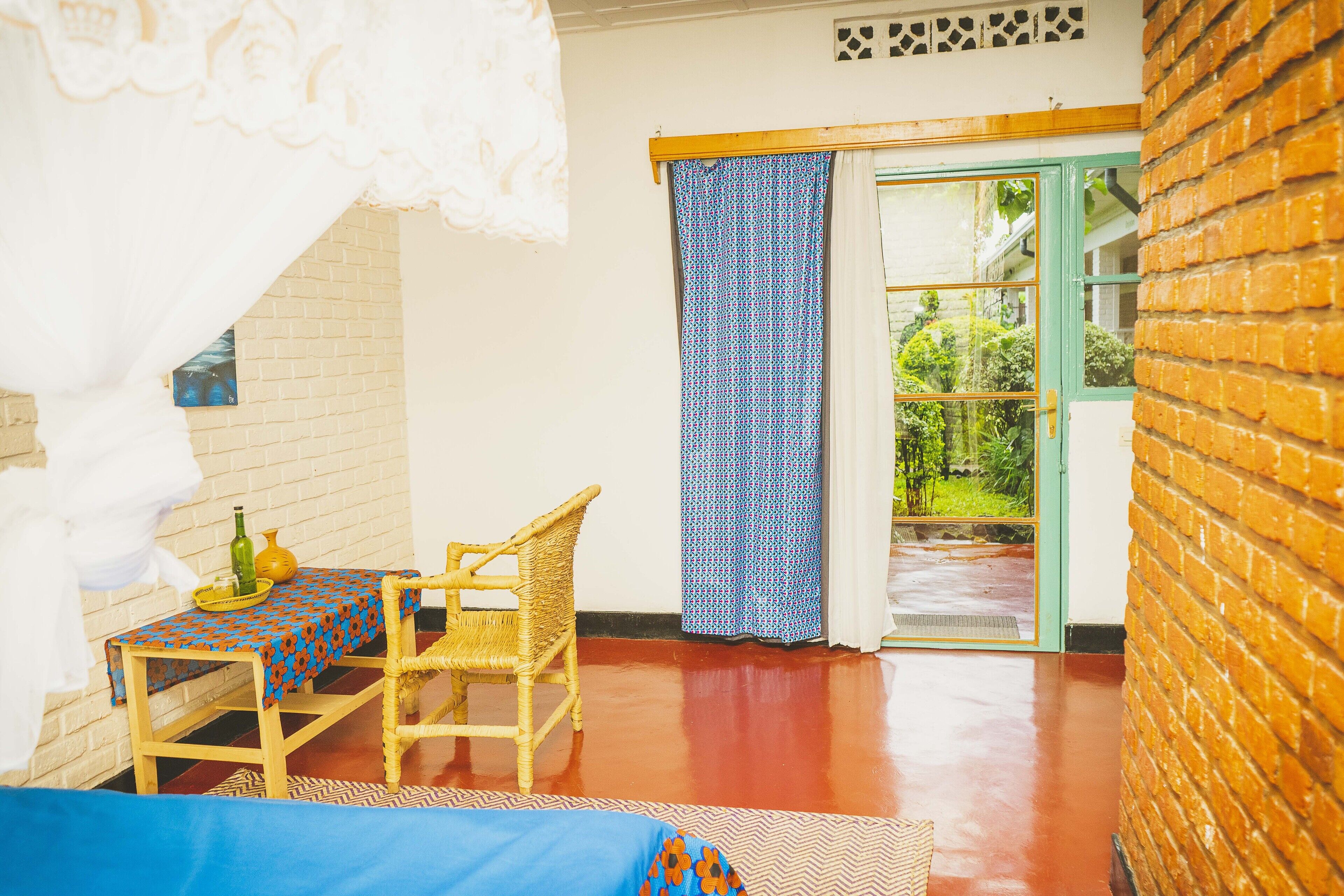 A room with a brick wall, a wooden table with a patterned tablecloth, a wicker chair, and a view through a glass door to an outdoor area.