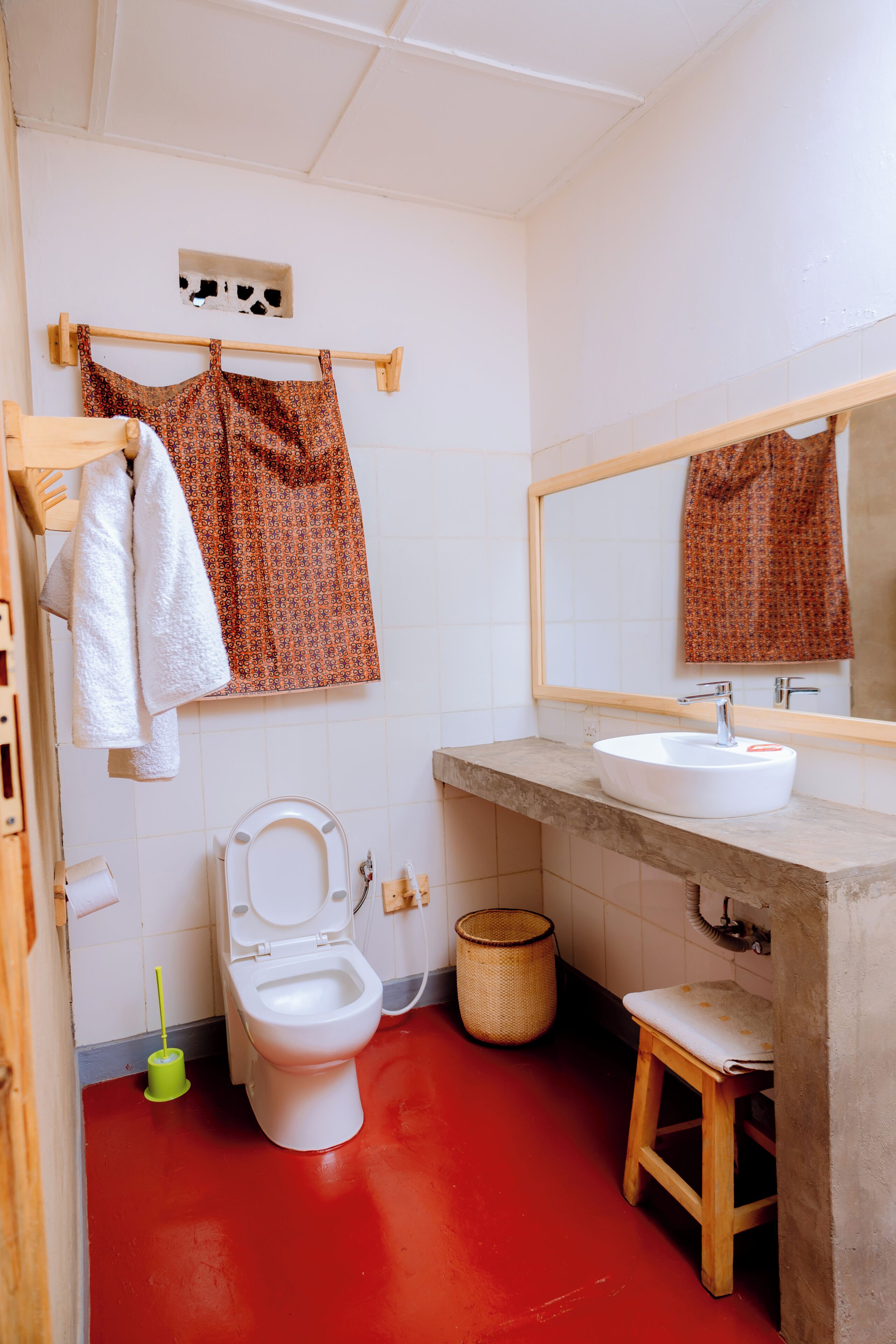 A bathroom with a white toilet, a sink, a mirror, and a red floor. There are two towels hanging on a rack and a basket on the floor.