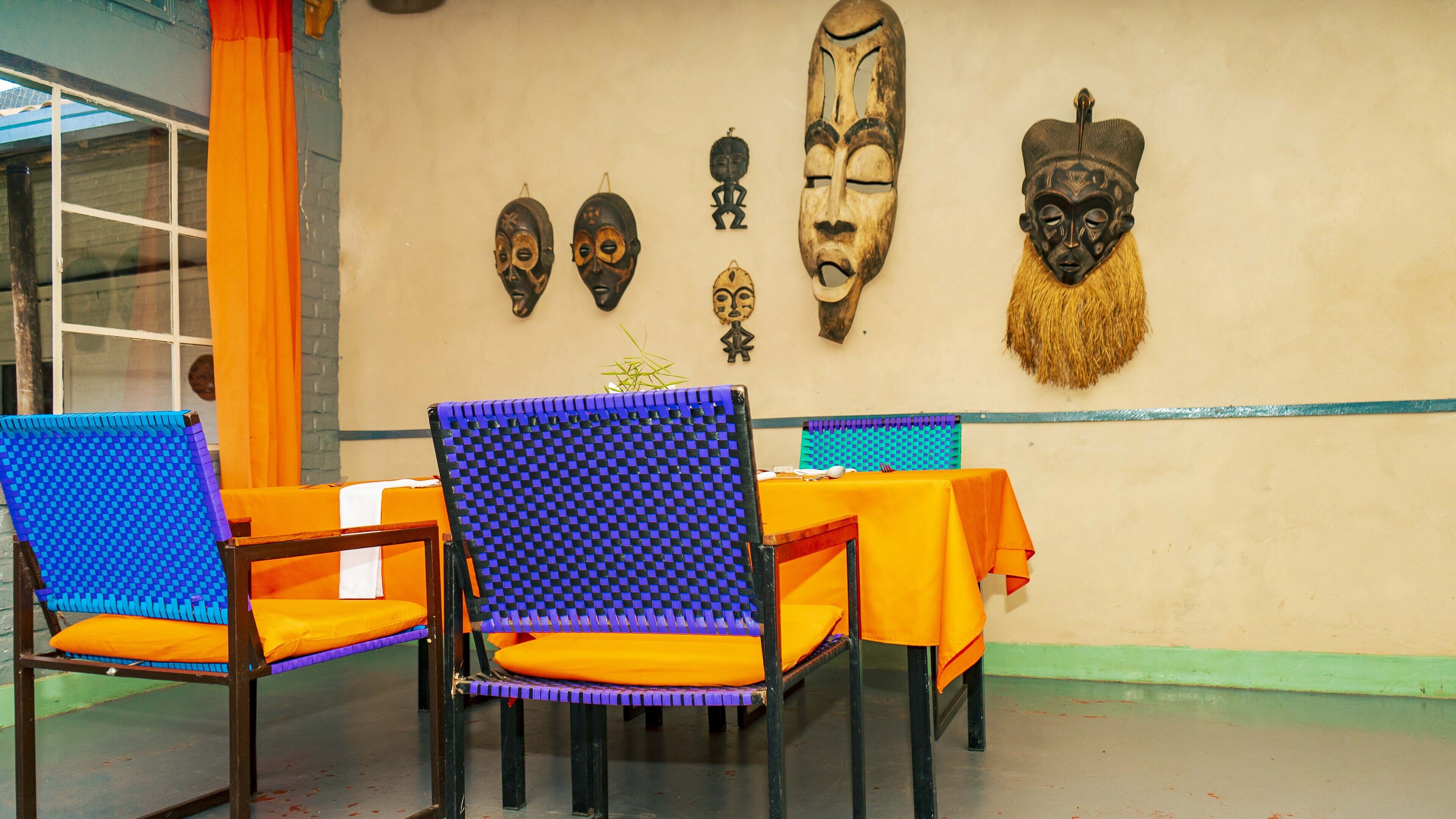 Un coin repas avec deux chaises bleues et orange, une table recouverte d’une nappe orange et plusieurs masques en bois accrochés au mur.