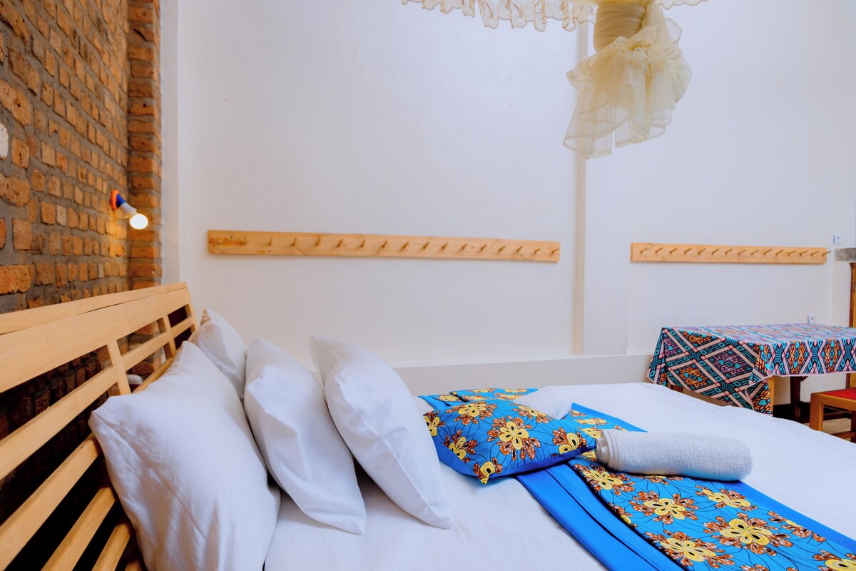 Habitación de hotel con pared de ladrillo, cabecera de madera, cama con ropa de cama blanca y azul, y una mesa con mantel estampado.