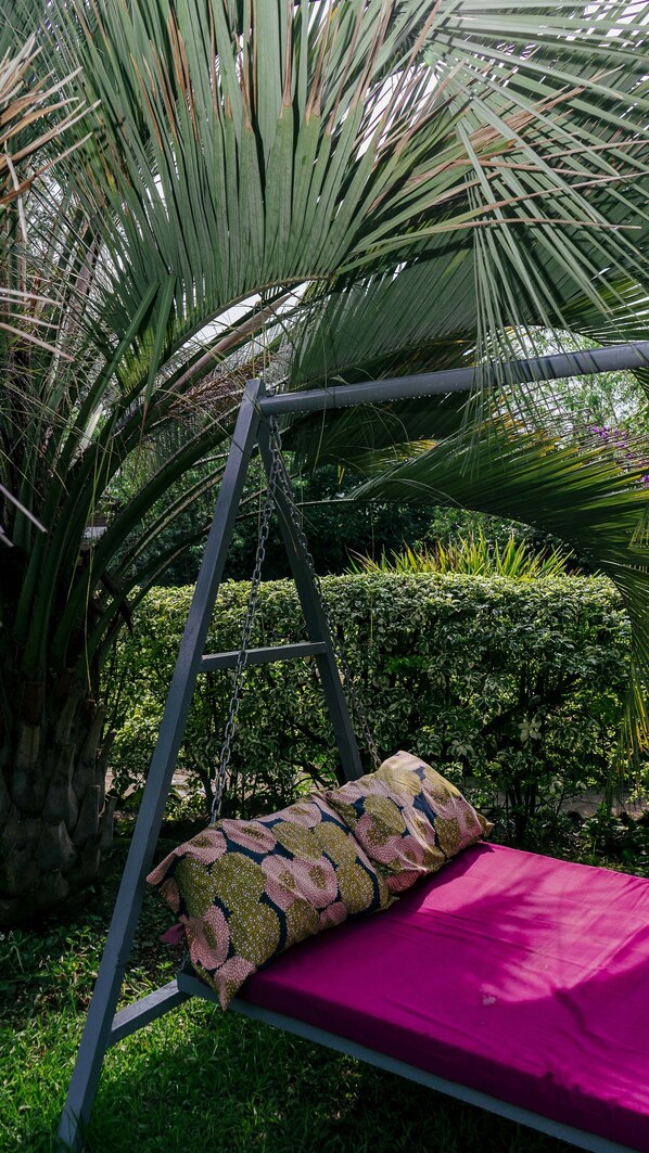 Una silla colgante con un cojín estampado y un colchón rosa, frente a un fondo de vegetación exuberante y una palmera.