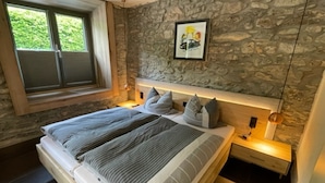 A bedroom with a stone wall, a large bed with grey bedding, a window with curtains, and a framed picture on the wall.