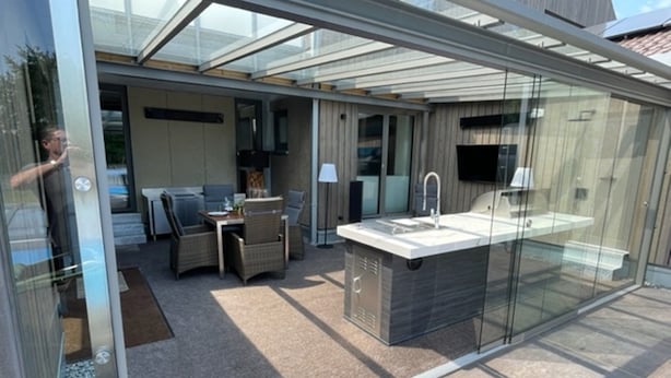 A modern outdoor kitchen area with a glass enclosure, a dining set, and a built-in sink.