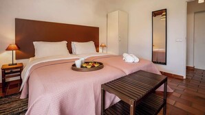 Une chambre d’hôtel avec un grand lit, une table de chevet avec une lampe, un plateau de fruits, un banc en bois, une armoire et un miroir au mur.