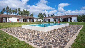 Un espace piscine doté d’un sol en pierre, entouré de bâtiments blancs aux toits couverts de tuiles, et un ciel d’un bleu limpide.