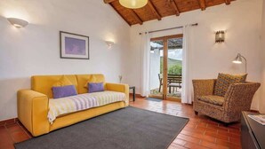 A cozy living room with a yellow sofa, a wicker chair, and a view of the outdoors through a glass door. - Burro Ville - Serene Houses w/ Pool by HostWise (Portimão)