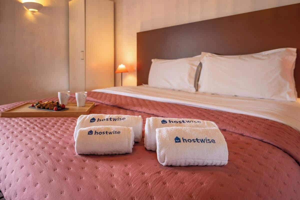 A hotel room with a bed, pillows, a bedside table with a lamp, and towels with the 'hostwise' logo.
