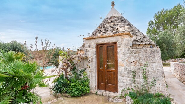 Una cabaña de piedra con techo cónico y puerta de madera, rodeada de vegetación y cielo despejado.