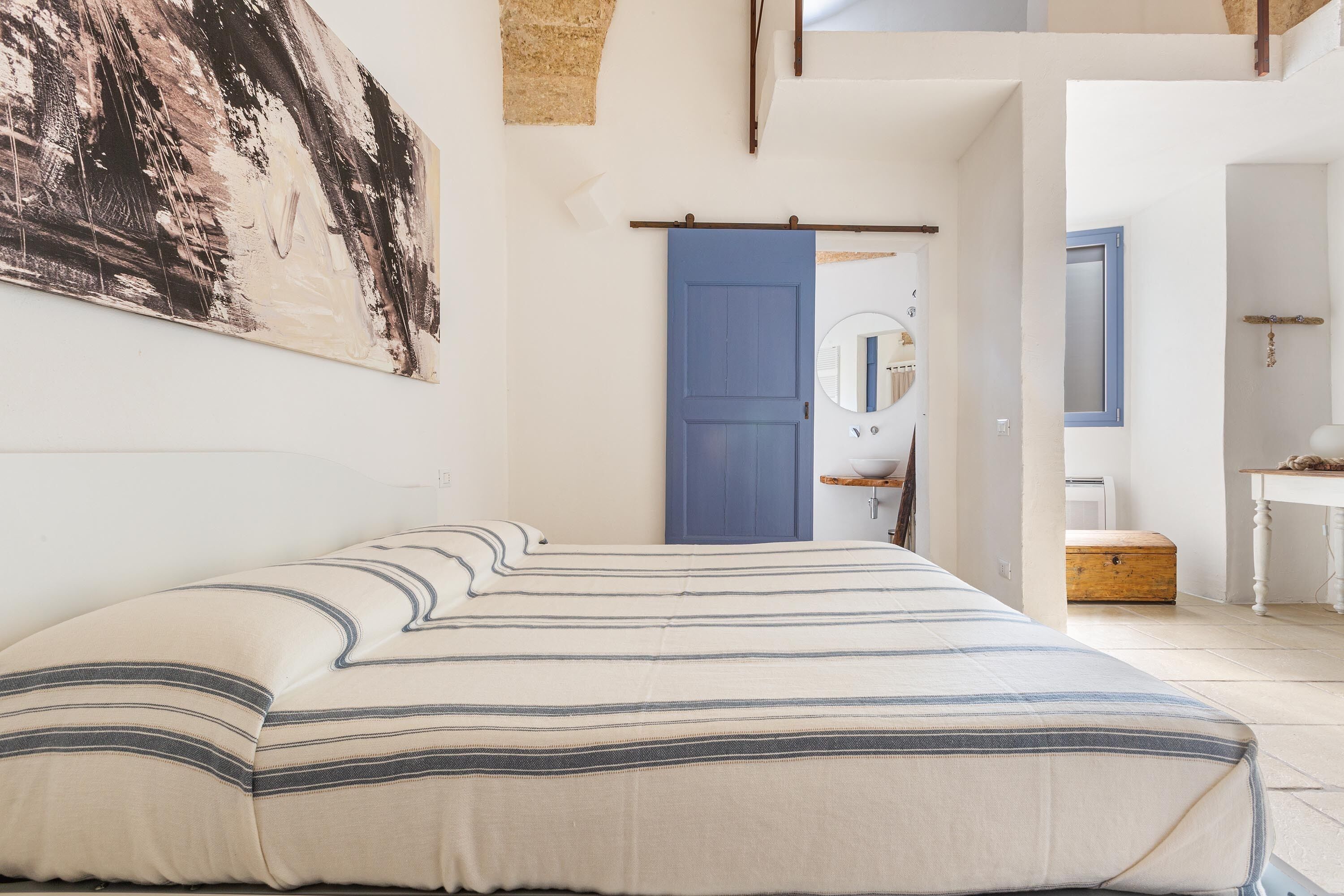 Ein Schlafzimmer mit einem Bett, einer blauen Tür, einem Spiegel und einer Holzkiste.