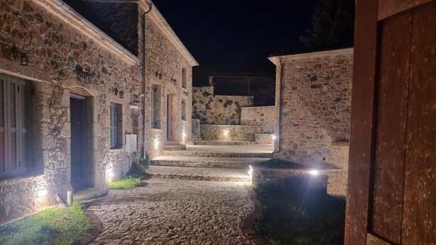 Un sendero de piedra que conduce a un edificio con puerta de madera, rodeado de muros de piedra e iluminado por luces en el suelo.