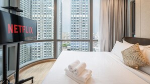 A hotel room with a large window, a bed with white linens, a flat-screen TV, and a view of high-rise buildings. - SaigonHomes in Vinhomes Central Park (Ho Chi Minh City)