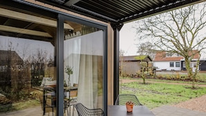 A modern outdoor patio with a glass door leading to a garden, a table, and two chairs. - Description (Waarde)