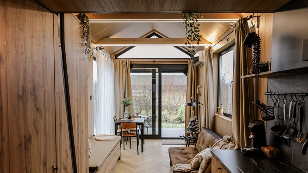 A cozy interior with a wooden floor, a sofa, a dining area, and a kitchen with hanging utensils. - Description (Waarde)