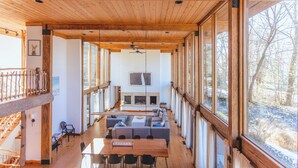 A spacious interior with a high ceiling, wooden beams, a fireplace, and large windows offering a view of the outdoors.