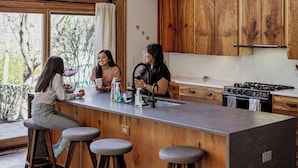 A modern kitchen with wooden cabinets, a large island with stools, and a view of trees outside.