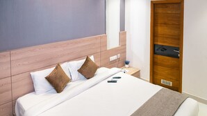 Een net opgemaakt bed met witte lakens en twee bruine kussens, een houten hoofdbord, een nachtkastje met een afstandsbediening, en een houten deur met een handvat.