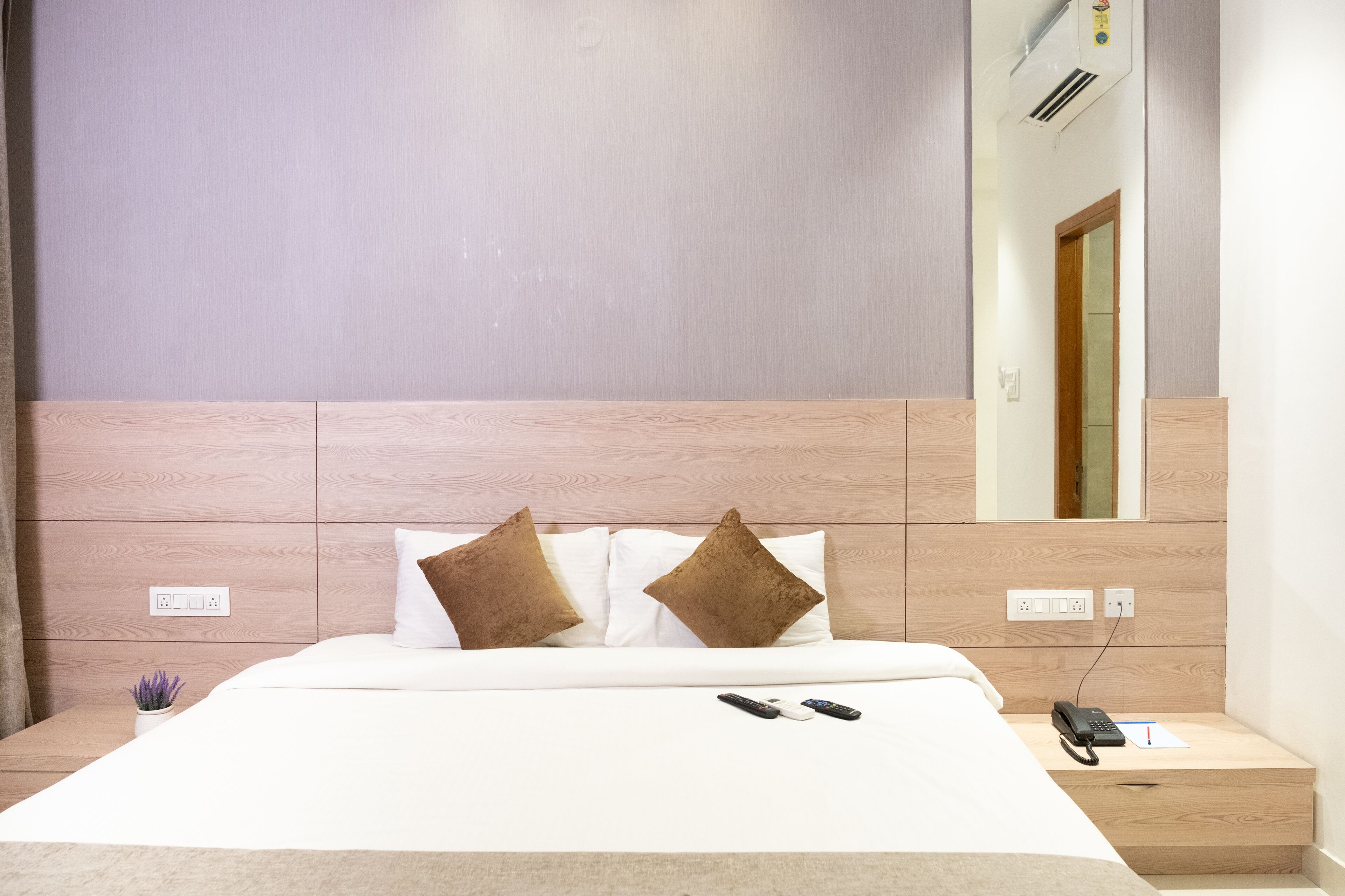 A neatly made bed with a white comforter, two brown pillows, and a bedside table with a phone and remote controls.